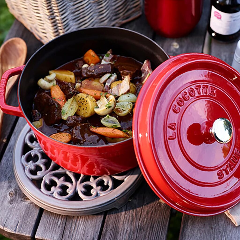 Red cast iron Dutch oven with a lid, containing beef stew with vegetables, on a gray trivet, on a wooden table.